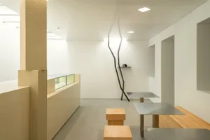 Empty restaurant interior with white walls, wooden tables, and chairs, featuring a potted plant and a clean, inviting atmosphere