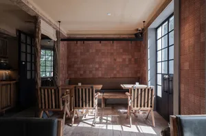 Rustic restaurant interior with wooden floors, tables, and chairs, and a brick wall, well lit with natural sunlight