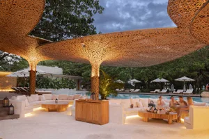 A resort patio with a wooden bar, lounge chairs, dining tables, umbrellas, and a swimming pool, set in a relaxing and inviting atmosphere