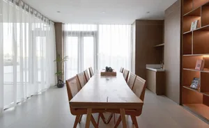 Open living space with a wooden dining table, chairs, and a kitchen area with a sink and cabinets, surrounded by natural light