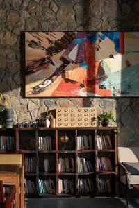 A cozy living area with a mixed media wall art, bookshelf, and two potted plants, creating a warm and inviting atmosphere