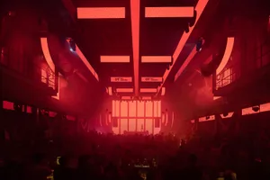A crowded indoor event venue with a red glow, people standing and socializing, and scattered chairs