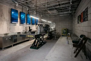 Industrial kitchen with concrete floors and walls, featuring a countertop with various items and chairs, lit by overhead lighting