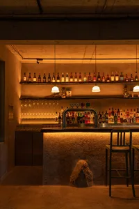 A cozy bar area with a long wooden counter featuring bottles and wine glasses, accompanied by two chairs in a warm and inviting atmosphere