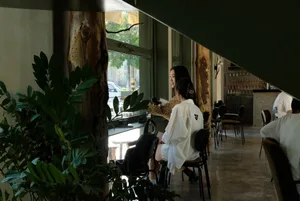 Two women sit at a dining table surrounded by wine glasses, cups, bowls, vases, and bottles in a relaxed restaurant setting