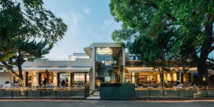 A modern restaurant with a green building, surrounded by trees, featuring dining tables, chairs, and potted plants, conveying a warm and welcoming ambiance