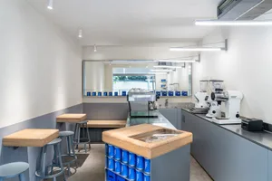 Industrial kitchen with a dining table and chairs, featuring coffee makers, cups, and bottles, in a well-equipped and functional space