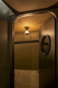 A dimly lit hallway with a black door, a round window above the doorway, and two light fixtures, one near the center and one to the right