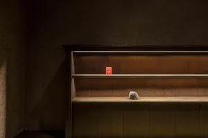 A wooden bookshelf in a darkened room with a small red object and a rock on one of the shelves