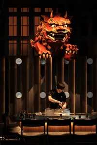 A professional chef prepares food in an Asian restaurant kitchen with a dragon head statue, surrounded by bowls and bottles, in a focused and professional atmosphere