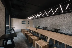 A modern restaurant interior with a large wooden dining table, black ceiling, and hanging lights, surrounded by chairs and potted plants