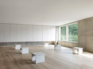 Open living area with a large white wall, two windows, concrete walls, wooden floor, and natural light