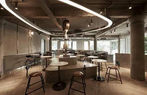 A modern dining space with a large circular table, smaller tables, chairs, concrete walls, and wooden accents, lit by overhead lamps