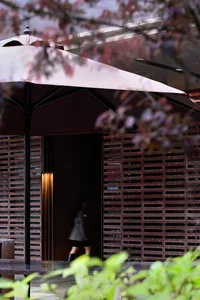 A woman in a black dress walks through a doorway with two umbrellas and a chair in a residential or commercial space with outdoor elements