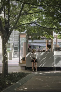 A casual outdoor cafe scene with a woman standing in front of a small white food stand, surrounded by potted plants and other people in the background