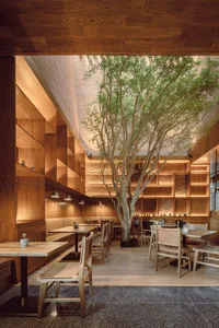 Warm and inviting restaurant interior with wooden floors, tables, chairs, and a central tree, offering multiple seating options