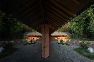 A woman walks towards the entrance of a traditional Asian building with a brick wall, wooden roof, and bamboo elements
