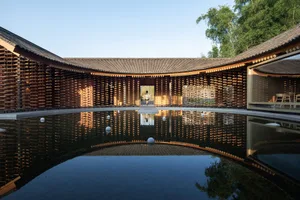 A serene Asian-style building with a wooden roof surrounded by trees, featuring a courtyard with a pond and several people in a peaceful environment