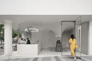 A modern cafe interior with white walls, glass door, and natural light from a large window, featuring chairs and dining tables, with two people inside looking at something