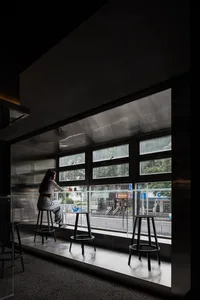 A woman sits at a counter in a cafe with a glass window, surrounded by chairs and a dining table, in a calm and relaxed atmosphere