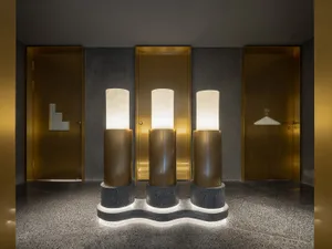 A grand hallway with three tall gold marble columns lit by side lights, set against a gray stone floor