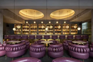 A modern bar area with purple chairs and tables, surrounded by dining tables and a well-equipped counter with numerous bottles on display