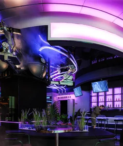 A modern bar area with a purple ambiance, featuring a circular table, chairs, potted plants, and a sleek bar with stainless steel or contemporary materials