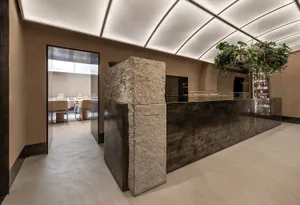 A restaurant interior with a stone wall separating two dining areas, featuring chairs, tables, and potted plants in a relaxed atmosphere