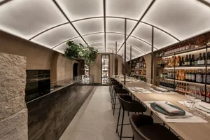 Modern restaurant interior with a large dining area, wooden furniture, and a bar section, featuring wine bottles and warm lighting