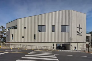 A large commercial building with a white exterior, multiple windows, and a modern appearance, set against a backdrop of parked cars and an urban landscape
