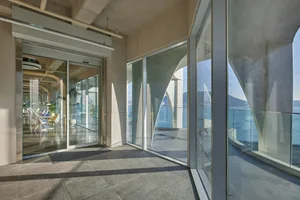 Open living area with concrete flooring, glass walls, and natural light from large windows, featuring two chairs and a potted plant