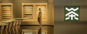 A woman stands in a large room with decorative cups and vases on display, against a backdrop of various items on the walls