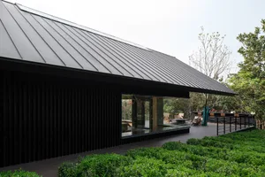 A modern house with a black roof and green facade features a large window and outdoor seating area with chairs and a bench, set in a calm and inviting atmosphere