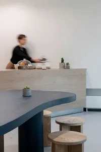 Warm and inviting cafe space with wooden floors and walls, featuring chairs, a dining table, and potted plants, with a woman in the background