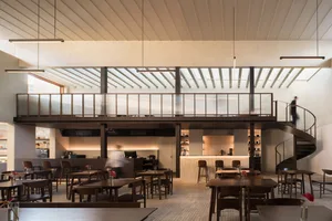 A modern restaurant interior with a large dining area featuring wooden floors, white walls, and well-lit tables and chairs, with patrons seated and standing