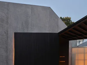 A modern building with a gray wall and a roof, featuring a black doorway with a sign above it, set against a backdrop of a tree