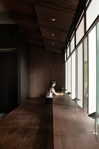 Woman sitting at a wooden dining table in a dimly lit room with a vase and a bench