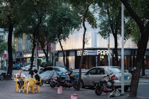 城市街景，白色建築、停放汽車、機車與行人來往，椅凳散置各處