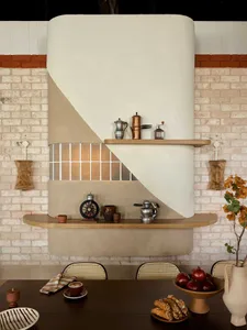A warm and inviting kitchen space with a brick wall, white countertop, and various kitchen items scattered around, including chairs, cups, bowls, apples, and bottles, with a clock on the wall
