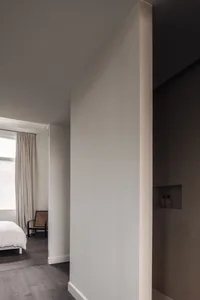A dimly lit bedroom with a white bed and two chairs, surrounded by white walls and a minimalist aesthetic