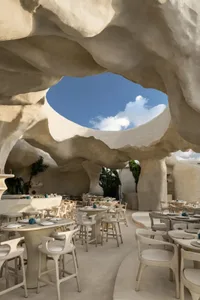 A spacious restaurant interior with white stone walls, multiple tables, chairs, and potted plants, creating a clean and inviting atmosphere