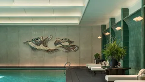A large indoor pool area with a mosaic tile design on the walls, surrounded by seating and potted plants, creating a tranquil atmosphere