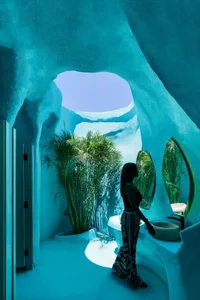 A modern bathroom scene with a woman standing in front of a mirror, a chair, a bowl, and a sink, with blue walls and a mix of natural and artificial lighting
