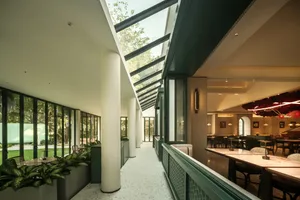 Modern restaurant interior with green walls, glass ceiling, and natural light, featuring dining tables and chairs, and potted plants
