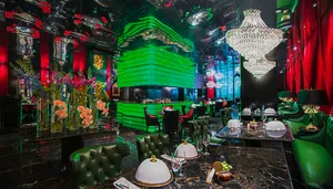 A modern restaurant with a large dining area featuring multiple tables, chairs, wine glasses, cups, and bowls, set against a backdrop of green walls