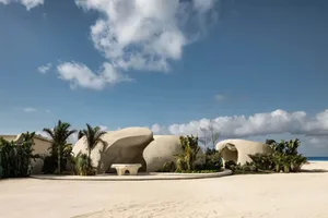 A beachfront resort with a large white building surrounded by smaller structures, palm trees, and sand, featuring modern architectural design