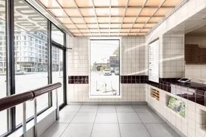 A modern interior space with a tiled floor, walls, and a large window allowing sunlight to enter, featuring two benches and a view of a car outside
