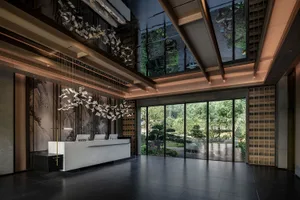 A modern lobby with a large window, glass doors, a white countertop, a wooden desk, and several chairs, with three potted plants adding greenery to the space