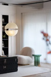 A modern living area with a white chair in front of a glass table, surrounded by books and a suitcase, filled with natural light from a large window