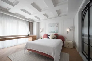 A calm and inviting bedroom with a large white bed, pink headboard and footboard, two nightstands with lamps, a couch, and a chair